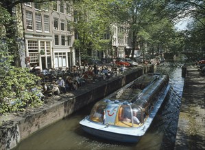 HOLLAND, Noord, Amsterdam, Glass roofed tourist boat passing canal side cafe on the Lelegracht