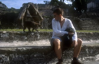 GUATEMALA, Tikal, Tourist smoking a cigarette with Coati walking along the wall behind in the main