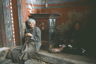 BHUTAN, Kyicho Temple, Man and elderly woman sitting on the floor beside a prayer wheel on a plinth