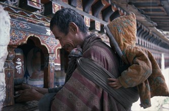 BHUTAN, Kyicho Temple, Man with child on his back placing butter on a prayer wheel as an offering