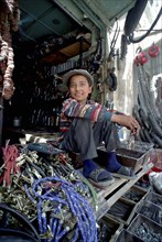 CHINA, Xinjiang, Kashgar, Young hardware vendor sitting amongst goods for sale