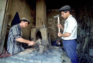 CHINA, Xinjiang, Kashgar, Blacksmith beating heated metal in his workshop with helper