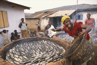 GHANA, Near Accra, "Smiling woman smoking fish in large, circular clay oven."