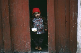 CHINA, Guangxi, Yangshuo, Child holding bowl of rice and chopsticks looking out from narrow doorway