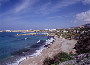 CYPRUS, Coral Bay, Man walking along shore of quiet beach with small harbour behind lined by palms