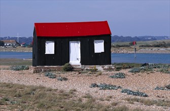20059206 ENGLAND East sussex Rye Rye harbour. Red and black corrugated hut with white doors built on the shingle with the harbour inlet behind.