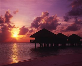 MALDIVES, Baros Island, View of silhouetted beach huts on stilts over water against a dramatic pink