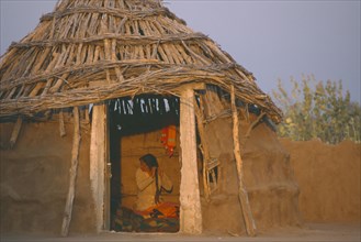 INDIA, Rajasthan, Thar Desert, Bhikodai.  Early morning light on thatched mud hut with woman