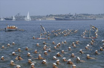 RUSSIA, Sevastopol, Navy Day. Sailors swimming in the sea in formation with battleships behind.