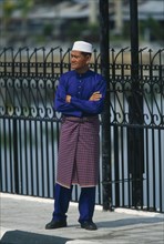 BRUNEI, People, Men, Man standing on pavement with arms crossed next to railings dressed up to