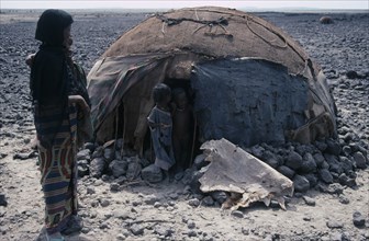 ETHIOPIA, Danakil Depression, Afar woman carrying child standing outside hut or ari with two young