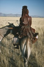 NAMIBIA, Marienfluss, Himba tribeswoman on donkey seen from behind wearing typical leatherwork
