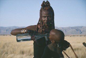 NAMIBIA, Marienfluss, Himba woman giving child a drink of water from Coca Cola bottle.  The Himba