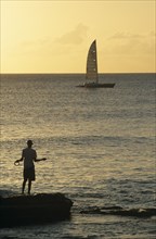 WEST INDIES, Barbados, Bridgetown, Fisherman silhouetted in the evening light with yacht on the