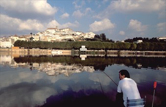 PORTUGAL, Coimbra, Man fishing on the bank of the River Mondego with the University crowning