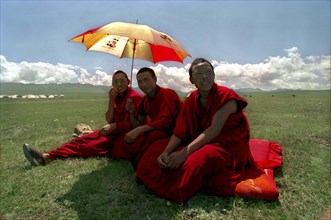 CHINA, Gansu, Xiahe, Three monks with umbrella sitting in green open landscape watching festival