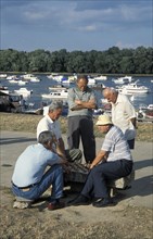 SERBIA, Belgrade, Group of men gathered around playing Chess with boats on The Danube river behind.