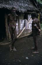 SOLOMAN ISLANDS, Indigenous People, Two men wearing penis sheaths standing outside thatched hut.