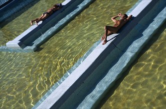 HUNGARY, Budapest, Man and woman lying on raised tiled area above water level in spa.