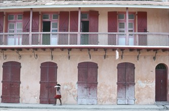 HAITI, Cap Haitien, Street scene with woman walking past building with faded pink plaster walls and