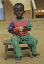 GHANA, Children, Portrait of young child sitting on wooden stool holding packet of chocolate.