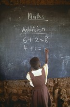 GHANA, West, Education, Child in primary school maths class writing sums on blackboard with chalk.