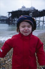 CHILDREN, Playing, Toddler dressed in red coat crying on Brighton Beach with view of the crumbling