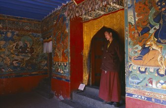 TIBET, Sakya, Monk standing in a doorway with brightly painted religious scenes and deities around