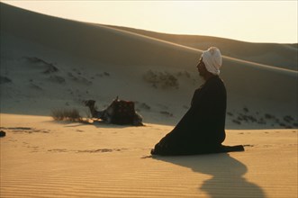 EGYPT, Religion, Muslim man praying in the desert with camel and sand dunes behind.