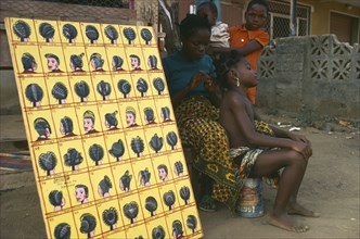 NIGERIA, Lagos, Hairdresser braiding hair of young girl at roadside beside board displaying