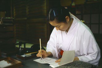 JAPAN, Honshu, Tokyo, Meiji Jingu Shrine.  Japanese woman writing at desk using brush and ink.