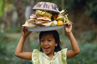 INDONESIA, Bali, Laughing Balinese girl carrying food offerings on her head.