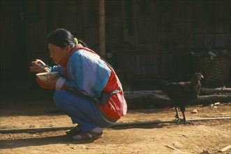 THAILAND, Chiang Rai Province, Doi Lan, Lisu woman eating whilst squatting outside her home with a