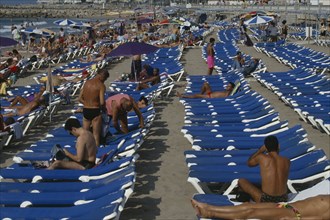 SPAIN, Catalonia, Sitges, Crowded beach with rows of blue sun loungers and sunbathers.