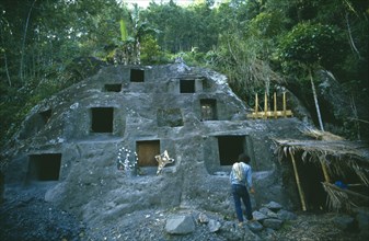 INDONESIA, Toraja, Stone burial ground surrounded by trees with man in the foreground.