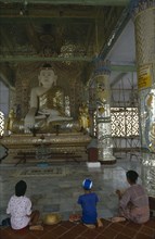 MYANMAR, Sagaing, Woman and children kneeling in front of giant seated Buddhist figure.