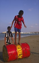 OMAN, Sur, Two young Omani boys playing on an abandoned Shell oil drum on the beach.