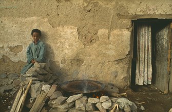 ERITREA, Asmara, Child sitting outside poor household with injera cooking on open fire at side.