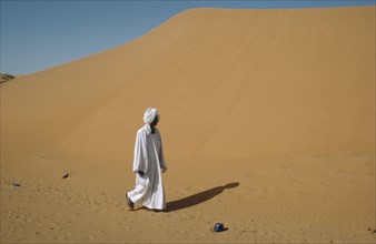 SUDAN, Kordofan Province, El Humra Village, Man in white robe and turban passing huge sand dunes