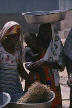 SUDAN, Work, Nigerian woman winnowing grain beside woman carrying child in her arms and large dish
