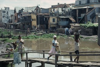 MADAGASCAR, Antananarivo, Children crossing wooden bridge over stagnant water in poor housing area