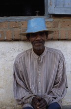 MADAGASCAR, Ihosy, Portrait of an elderly man sitting against a wall wearing a blue straw hat