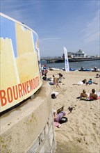 ENGLAND, Dorset, Bournemouth, Tourists on the sand of the West Beach beside the pier. A sign