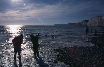 ENGLAND, East Sussex, Birling Gap, People in silhouette paddling at the waters edge on pebbled
