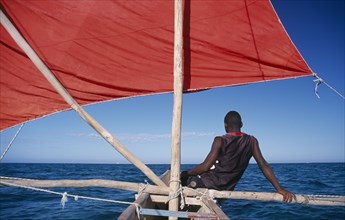 MADAGASCAR, Tulear, Ifaty Beach. Young fisherman sitting on bow of red sail Pirogue boat