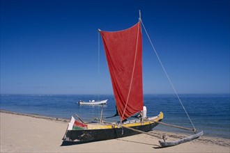 MADAGASCAR, Tulear, Ifaty Beach. Pirogue boat with red sail on sandy beach next to the waters edge