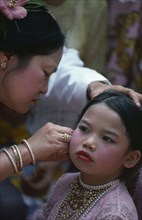MYANMAR, Children, Young girl wearing make up and jewellery having her ear pierced by older woman.