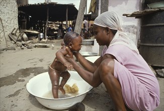 GHANA, Dixcove, Young mother washing baby in bowl in street.