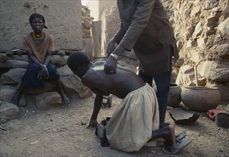 MALI, Tribal People, Chiropractor manipulating client in Dogon village with woman sitting at side.