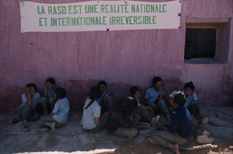 WESTERN SAHARA, SADR, Sahrawi schoolgirls sitting against pink plastered wall under banner with